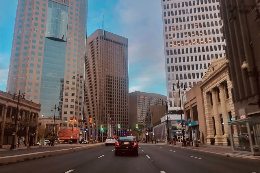 Cars are driving at the intersection of Portage and Main in downtown Winnipeg. Highrise buildings are in the background.