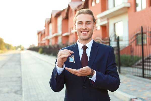 A smiling male real estate agent in a suit holds the keys to a rental unit. Using a realtor to find a rental is a smart strategy for newcomers to Canada