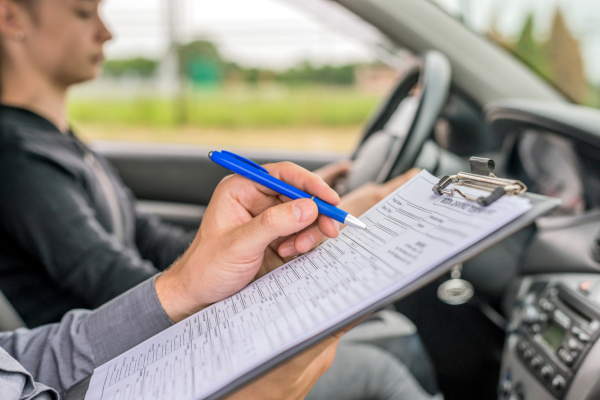 A young woman is completing a driving test. A driving instructor is writing on a clipboard.