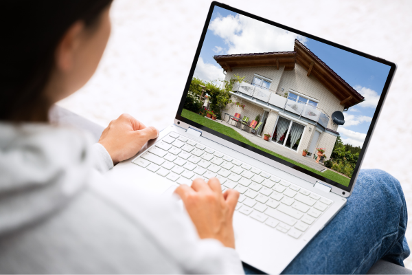 A woman in blue jeans and a hoodie looks at a house in the screen of her laptop. October's housing market report shows sales on the rise