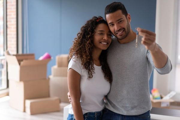 A smiling young couple stand in the living room of a home they just bought holding the keys. Canada's housing market report says sales rose in October