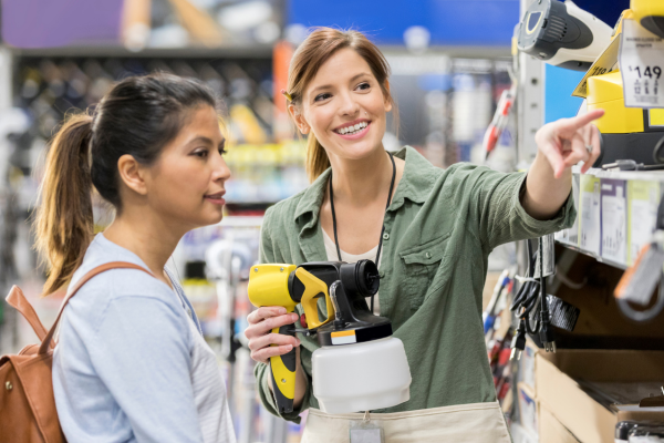 A female retail sales clerk holding a paint spray gun points to a price sticker for a female customer with a ponytail.