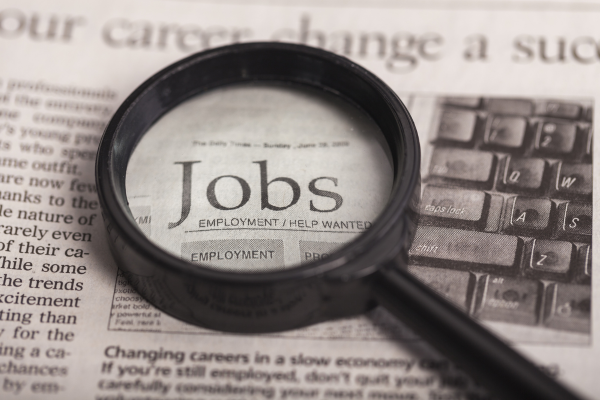 A magnifying glass lying on a help wanted page in a newspaper magnifies the word "Jobs." The latest job market report shows that Canada is still adding jobs every month.