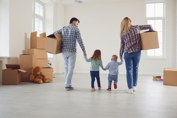 A man and a woman with their backs to us and each carrying a box hold the hands of their two young children inside a home