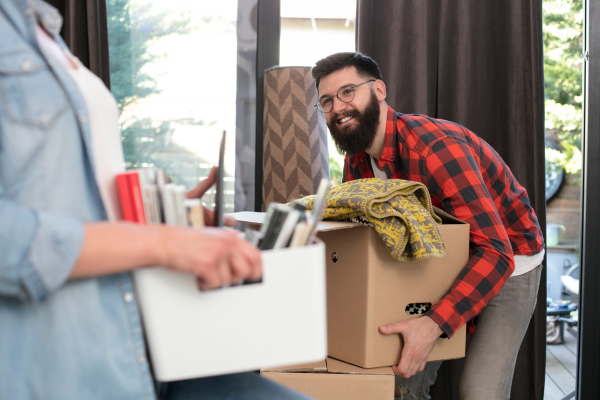 A bearded man in a red plaid shirt lifts a box as a woman holding a box looks on as they move into their apartment.