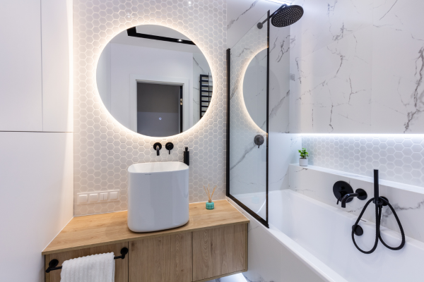 A modern bathroom featuring an illuminated round mirror overtop of a raised sink and beside a glass shower.