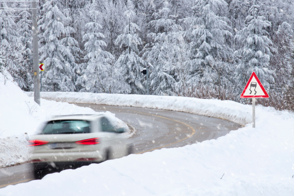 A car is driving on an icy road. The car is approaching a road sign that indicates icy and slippery road conditions. 