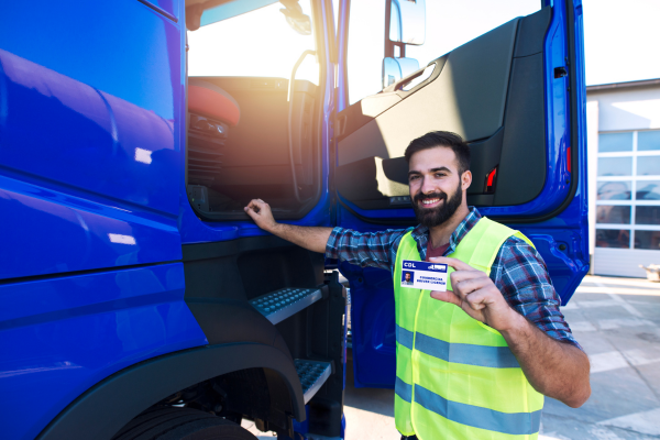 A truck driver stands by his truck holding a Commercial Driving License